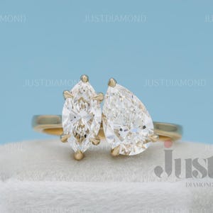May include: A gold ring with two pear-cut diamonds. The diamonds are positioned next to each other, showcasing a sparkling cut. The ring is displayed on a white surface, set against a light blue backdrop.