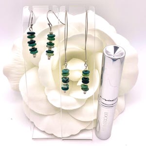 May include: An image showcases three pairs of earrings with a dangling design. Each earring features a series of small, round, green beads strung together. A silver lipstick tube is also visible. The earrings are displayed on a clear stand against a white background.