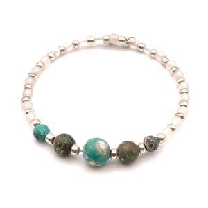 May include: A beaded bracelet featuring a mix of white, turquoise, and brown beads. The bracelet has a delicate design with small white beads and larger turquoise and brown accent beads. The beads are strung together with silver-colored metal.