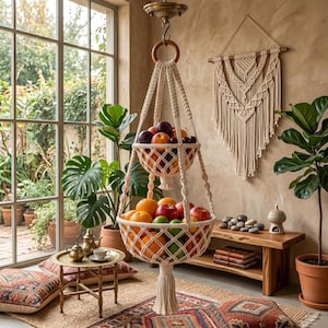 Puede incluir: Una cesta de frutas de macramé de dos niveles llena de manzanas, naranjas y ciruelas. La cesta de color crema cuelga de un anillo de madera y un gancho de techo. La habitación tiene un estilo bohemio con plantas, una alfombra y un tapiz de macramé.