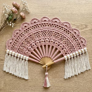 May include: A handmade, openwork crochet fan in a dusty rose color. The fan features a scalloped edge, a wooden handle, and cream-colored fringe and tassel accents. The fan is a decorative item.
