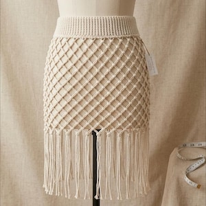 Pode incluir: Saia de macramé creme com um padrão de losangos tecido e detalhes de franjas. A saia é exibida em um manequim, mostrando sua textura e design. Uma etiqueta está presa à saia.