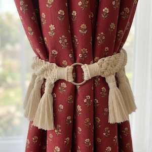 May include: Burgundy floral curtains held by a cream-colored macrame curtain tieback with a silver ring. The curtains have a repeating pattern of small, yellow and white flowers.