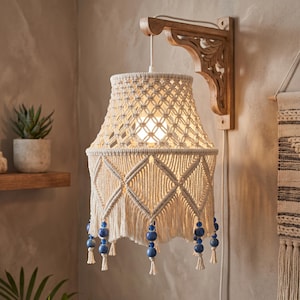 May include: A macrame lampshade with a cream-colored woven design, diamond patterns, and blue beaded accents. The lampshade hangs from a carved wooden wall sconce. The light is on, illuminating the intricate details of the macrame.