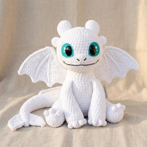 May include: A white crocheted dragon plush toy featuring large, bright teal eyes with black pupils, pointed ears, and wide, textured wings. The creature has a long, curved tail with a fin at the end and a small tag on its side.