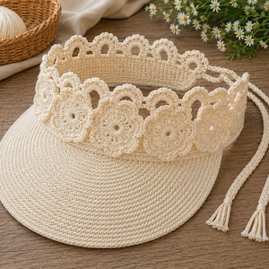May include: A cream-colored crochet sun visor featuring a wide, flat brim and a decorative crown band composed of interconnected floral motifs. The visor includes two long, braided ties with small tassels at the ends for securing the headpiece.