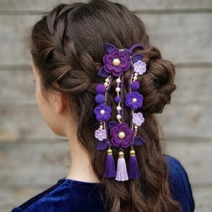May include: A handcrafted hair accessory featuring crochet flowers in shades of purple, adorned with pearls and tassels. The floral design includes various sizes of flowers and leaves, creating a decorative hairpiece.