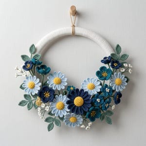 May include: A handmade crochet wreath with a white, wrapped base, decorated with blue and yellow flowers and green leaves. The wreath hangs from a wooden peg with a natural twine cord, offering a decorative touch for the home.
