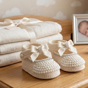 May include: A stack of folded cream-colored fabric tied with a satin ribbon, accompanied by a pair of cream-colored crocheted baby shoes featuring pearl embellishments, a central satin bow, and a rhinestone accent on the strap.