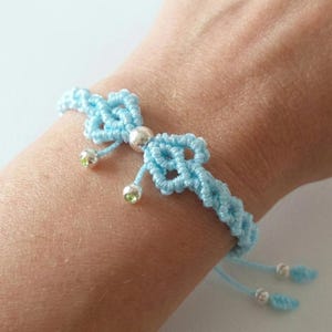 May include: A light blue macrame bracelet with a bow-shaped centerpiece and silver beads. The bracelet features a woven design and adjustable ties. Small green gems are also included in the design.