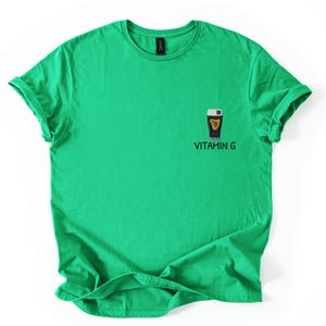 May include: A bright green t-shirt with rolled-up sleeves. The shirt features a small graphic of a dark pint of beer with a harp and shamrock, and the text "VITAMIN G" below the graphic.