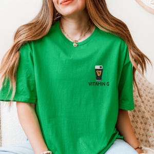 May include: A bright green t-shirt featuring a small graphic of a pint of stout with a shamrock and harp design. The text "VITAMIN G" is printed below the graphic in a sans-serif font. The shirt is a crew neck style.