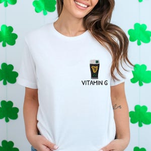 May include: White t-shirt with a small embroidered design of a pint of stout beer with a harp and shamrock. The text "VITAMIN G" is printed below the beer glass. Green shamrock decorations are in the background.