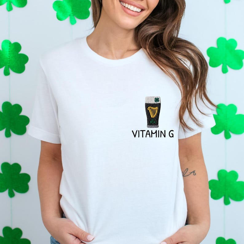 May include: White t-shirt with a small embroidered design of a pint of stout beer with a harp and shamrock. The text "VITAMIN G" is printed below the beer glass. Green shamrock decorations are in the background.