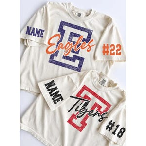 May include: Two off-white short-sleeve t-shirts. The top shirt features a large purple letter E, the orange script text Eagles, the word NAME on the sleeve, and #22. The bottom shirt has a red letter T, black script text Tigers, NAME, and #18.