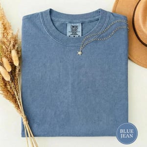 May include: A blue short-sleeved t-shirt with a Comfort Colors label at the inner neckline. A silver-toned chain necklace with a small star pendant rests on the fabric. The shirt is a solid blue color with a textured, washed appearance.