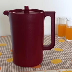 Può includere: Una caraffa in plastica color bordeaux scuro con coperchio abbinato e pomello centrale. La caraffa presenta un corpo cilindrico alto, un'ampia maniglia laterale e un piccolo beccuccio sul bordo superiore, progettata per servire bevande in cucina.