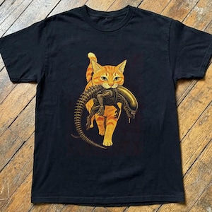 Pode incluir: Uma t-shirt preta de manga curta com uma impressão gráfica de um gato malhado laranja que carrega na boca uma criatura escura e segmentada com uma cauda longa e uma cabeça alongada. O gato é retratado a caminhar para a frente com foco.