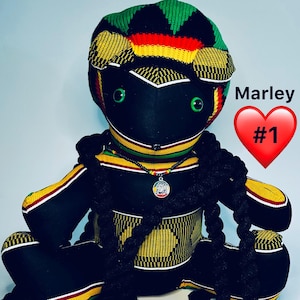 May include: A black doll with green eyes, wearing a Rastafarian-style hat in green, yellow, and red. The doll has long black braids and a necklace with a pendant. The doll is sitting with its legs crossed. The name "Marley" and a heart symbol are visible.