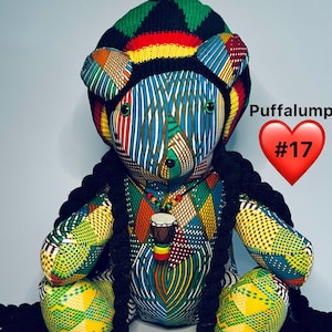 May include: A handmade teddy bear with a colorful patterned fabric design, wearing a knitted hat with red, yellow, green, and black stripes. The bear has black braids and a necklace with a small drum pendant. Text on the image reads "Puffalump #17" and "Bearjammin's".