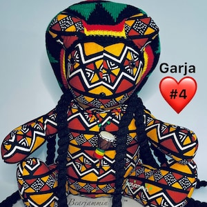 May include: A handmade teddy bear with a patterned fabric design in red, yellow, and black. The bear has long black braids and a matching hat. The text "Garja #4" is visible on the right side of the image. A small drum pendant hangs around the bear's neck.