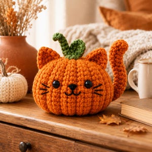 May include: A crocheted orange pumpkin shaped like a cat with two pointed ears, a green stem, a curved tail, black plastic eyes, and embroidered whiskers. The plush figure has a textured yarn surface and a rounded body sitting on a wooden surface.