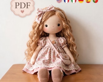 May include: A fabric doll with long wavy blonde hair, wearing a pink floral dress with a lace hem, a matching bow headband, and a small crossbody bag. The doll has black eyes and a small smile. A PDF label with a heart icon is visible.