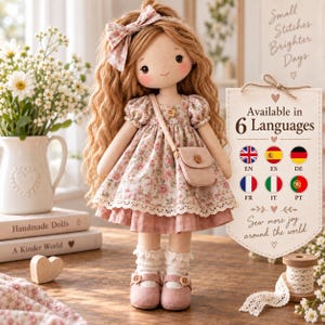 May include: A fabric doll with long wavy brown hair, a floral print dress, a matching hair bow, and a small crossbody bag. The doll wears white ruffled socks and pink shoes. Beside the doll is a sign with the text Available in 6 Languages.