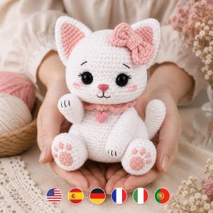 Puede incluir: Una figura de gato tejida a ganchillo en color blanco con un lazo rosa en la cabeza, un dije de corazón rosa en un collar festoneado y almohadillas rosas en las patas. El gato tiene grandes ojos negros, pestañas bordadas y nariz rosa.