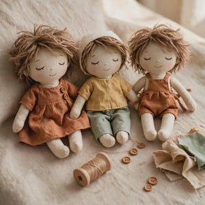 May include: Three handmade fabric dolls with embroidered closed eyes and rosy cheeks. The left doll wears a rust-coloured dress, the centre doll wears a yellow top and green trousers with a bonnet, and the right doll wears rust-coloured overalls.