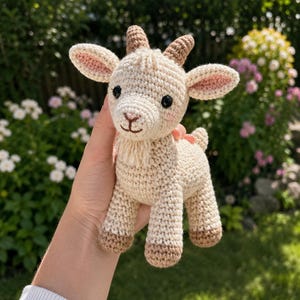 May include: A small, cream-coloured crocheted goat toy featuring two brown horns, black plastic eyes, a stitched nose and mouth, and a small beard. The goat has light pink blush on its cheeks and brown-coloured hooves on all four legs.
