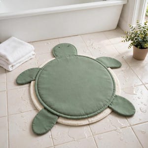 May include: A turtle-shaped floor mat featuring a sage green circular center with a matching head and four flippers, layered over a larger, round, cream-colored textured base. The mat is constructed with soft, padded fabric sections.