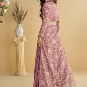 May include: A long, flowing mauve dress with a floral pattern and gold accents. The dress features long sleeves, a high collar, and a matching belt. The dress has a layered skirt with a ruffled hem.