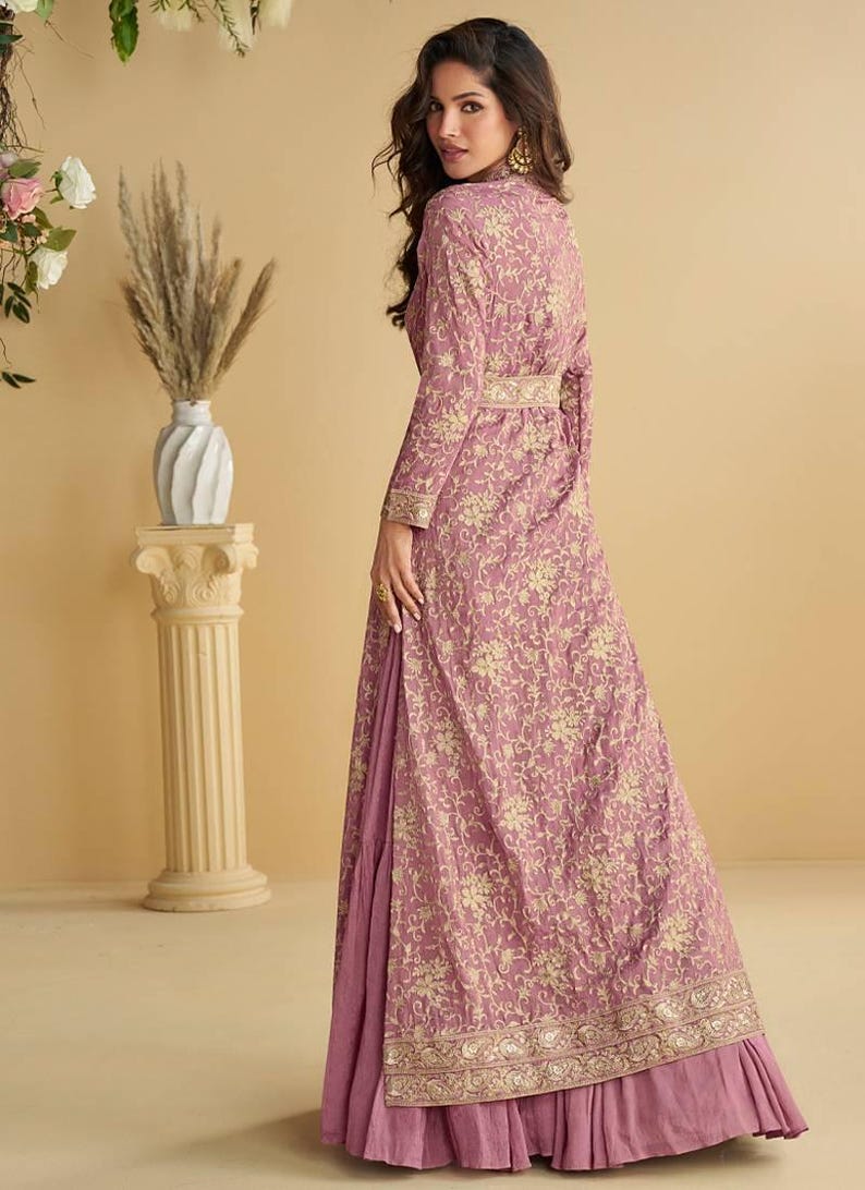 May include: A long, flowing mauve dress with a floral pattern and gold accents. The dress features long sleeves, a high collar, and a matching belt. The dress has a layered skirt with a ruffled hem.