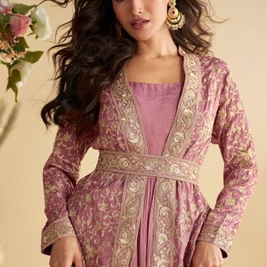 May include: A mauve-coloured, long-sleeved dress with intricate gold embroidery. The dress features a square neckline and a matching belt. The model is wearing gold earrings. The dress is suitable for formal occasions.