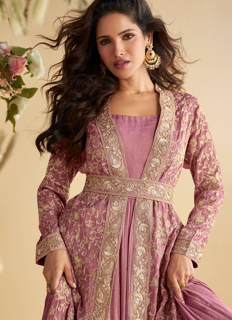 May include: A mauve-coloured, long-sleeved dress with intricate gold embroidery. The dress features a square neckline and a matching belt. The model is wearing gold earrings. The dress is suitable for formal occasions.