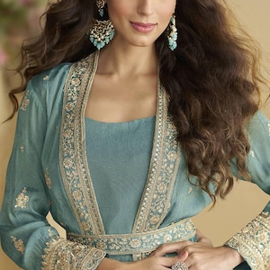 May include: A teal outfit comprising a long-sleeved jacket with detailed gold embroidery, worn over a matching dress. The ensemble is complemented by elaborate earrings and rings. The style is elegant and formal, suitable for special occasions.