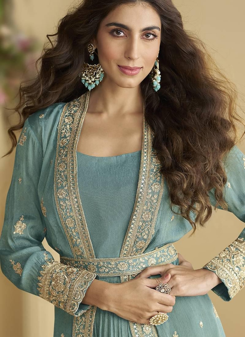 May include: A teal outfit comprising a long-sleeved jacket with detailed gold embroidery, worn over a matching dress. The ensemble is complemented by elaborate earrings and rings. The style is elegant and formal, suitable for special occasions.