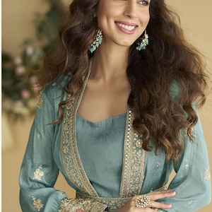 May include: A teal outfit comprising a long-sleeved top and a matching jacket, both adorned with gold embroidery. The ensemble is accessorized with a ring and earrings in a complementary gold and teal palette. The model has long, wavy brown hair.