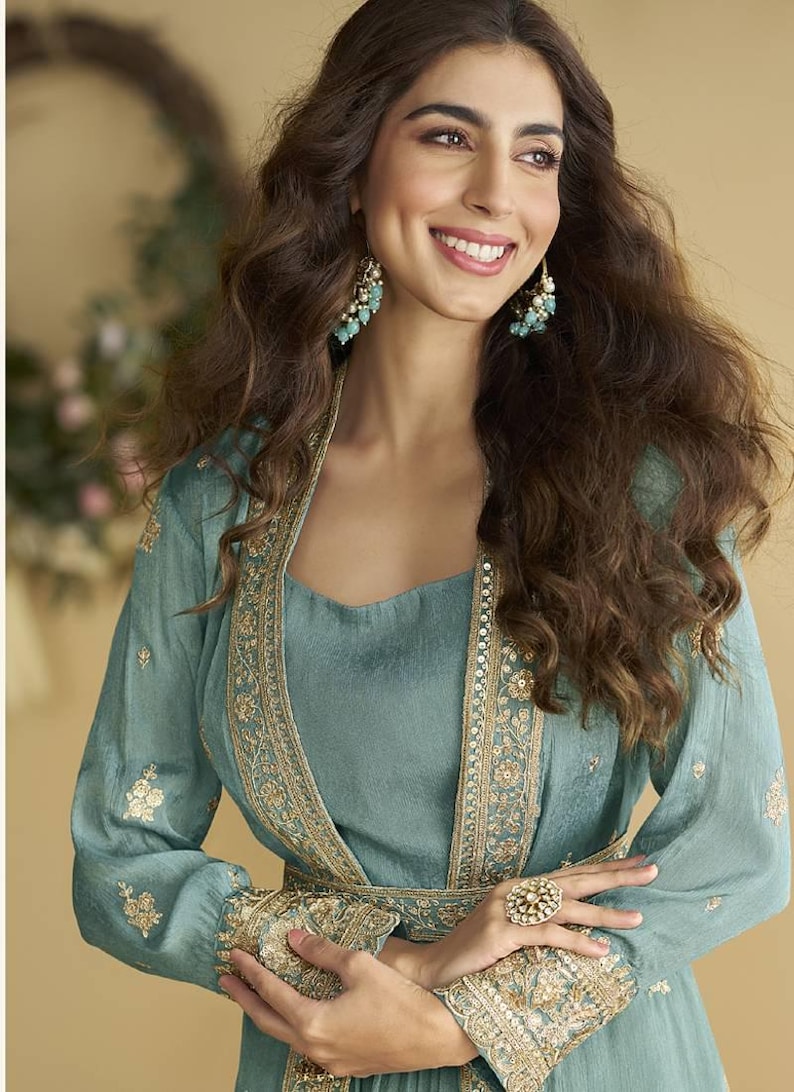 May include: A teal outfit comprising a long-sleeved top and a matching jacket, both adorned with gold embroidery. The ensemble is accessorized with a ring and earrings in a complementary gold and teal palette. The model has long, wavy brown hair.
