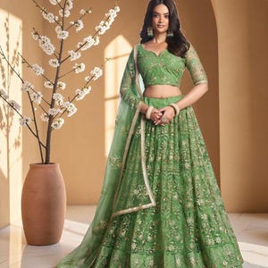 Puede incluir: Un lehenga verde bordado con un dupatta a juego. El atuendo presenta patrones florales dorados y se usa con una blusa verde a juego. La imagen también incluye un jarrón decorativo con flores blancas.