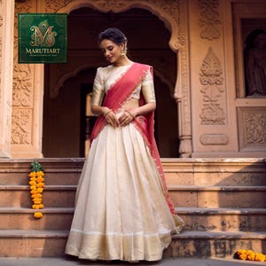 May include: A woman wearing a cream-colored skirt and blouse with gold accents and a red shawl. The skirt is pleated and has a gold border. The blouse has short sleeves. The background features an ornate building and the Marutiart logo.