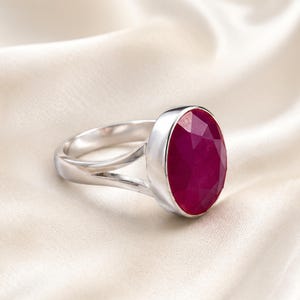 May include: A silver ring featuring a large, oval-shaped, faceted deep pink gemstone set in a smooth metal bezel. The band of the ring is polished silver and splits into two separate sections as it connects to the central gemstone setting.