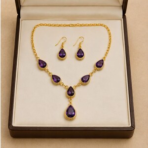 May include: A gold-toned jewellery set consisting of a necklace and a pair of matching earrings. The necklace features five teardrop-shaped purple stones set in gold frames, while the earrings each feature a single matching purple teardrop stone.