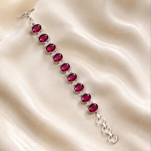 Peut inclure: Un bracelet en argent composé de dix pierres ovales à facettes d'un rouge profond, serties dans des chatons métalliques individuels. Le bijou présente un fermoir à barre à une extrémité et une série d'anneaux circulaires pour l'ajustement.