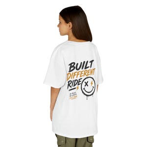 Könnte beinhalten: Ein weißes kurzärmeliges T-Shirt mit einem Grafikdruck auf der Rückseite, der den Text BUILT DIFFERENT RIDE in Schwarz und Gold, ein schwarzes Smiley-Gesicht mit X-Augen und den Text NO RULES NO LIMITS JUST RIDE in kleiner schwarzer und goldener Schrift zeigt.