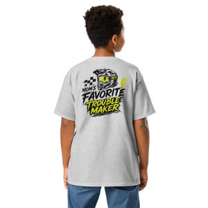 Könnte beinhalten: Ein hellgraues Kurzarm-T-Shirt mit einem Grafikdruck auf der Rückseite. Das Design zeigt einen schwarz-weißen Motocross-Helm mit gelber Schutzbrille, eine Zielflagge und den Text "MOM'S FAVORITE TROUBLE MAKER" in Schwarz und Gelb.