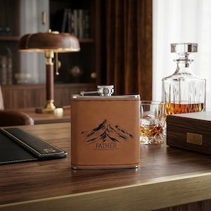 May include: A brown leather flask with a mountain graphic and the text "FATHER - Best Dad ever". The flask has a silver cap and is sitting on a wooden surface.