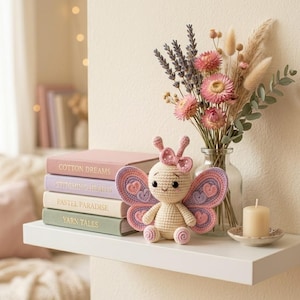 Può includere: Una bambola a farfalla all'uncinetto con ali rosa e viola è appoggiata su una mensola bianca accanto a una pila di libri. I libri hanno titoli come "Cotton Dreams" e "Stitching Hearts". Un vaso di fiori secchi e una candela accesa completano la scena.