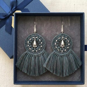 May include: A pair of handmade teal crochet earrings with silver-colored accents and fringe. The earrings are displayed in a navy blue gift box with a matching ribbon.